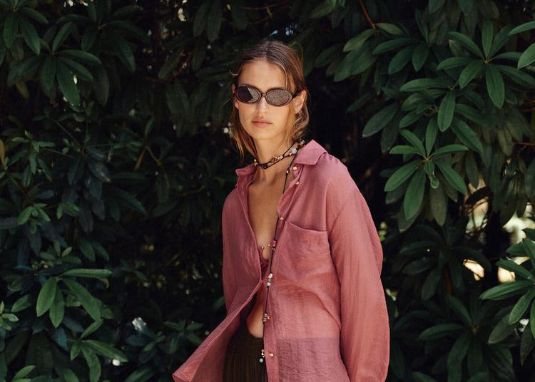 Person wearing a pink jacket and dark pants standing against a green leafy background